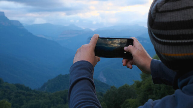 A man with a mobile phone in his hands takes pictures of a sunset in the mountains. A hiker takes pictures of a beautiful mountain sunset on his smartphone.