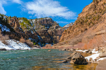 Fototapeta na wymiar View of Colorado river floating through Glenwood canyon in winter; Rocky Mountains and blue sky in background; distant view of state highway and moving red car