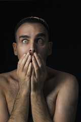 The man's eyes widened in surprise and fear. He covers his mouth with his hands. Portrait of a shirtless Caucasian man on a black background. A close-up. An emotional portrait of the man.