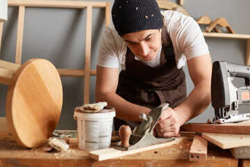 Portrait of handsome young adult man carpenter wearing white t-shirt, black cap and brown apron working in joinery, plane on the workbench in carpentry.