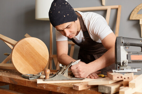 Image of young adult man carpenter wearing white t-shirt, black cap and brown apron working, planning wood in workshop, making wooden products, holding plane in hands.