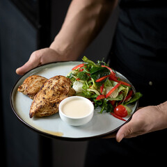 Baked chicken breast with fresh vegetable salad and sauce. Dish on white plate in hands of cook. Healthy diet. Fitness menu. Low calorie food. View from above. Soft focus. 