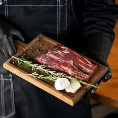 Raw piece of meat with rosemary and garlic on cutting board in hands of cook. Marble beef with spices. Cook in black overalls. View from above. Soft focus. 