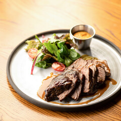 Steak with sauce and vegetable salad on plate. Meat is cut into pieces. Plate with dish on wooden table. Dinner at restaurant. Soft focus. View from above. Close-up.