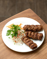 Lula kebab with side dish of fresh vegetables on round white plate. Asian cuisine, Oriental cuisine. Dish on white plate on wooden table. Pickled onions and carrots. Soft focus. View from above. 