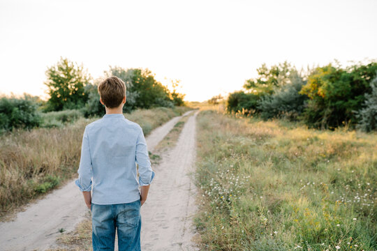 Teenager boy wearing blue shirt and jeans standing on the path in the field at summer, outdoor lifestyle photo. People from behind
