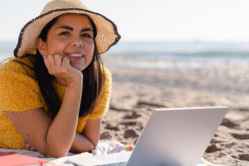 Latin freelance woman with hat working and enjoying a day at the beach lying in the sand. 