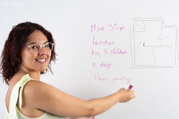 Candid young woman smiling, looking at the camera while explaining what is written on the white board with a pink marker.