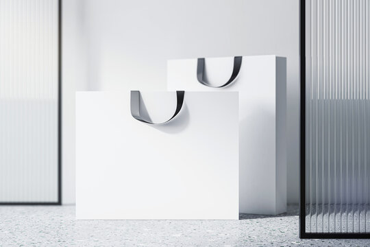 Front view on blank white paper shopping bags with space for your brand name or text on concrete floor near decorative glass partition and light wall background, close up. 3D rendering, mockup