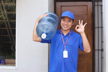 Office and home delivery, employees concept. Asian male courier in blue uniform, carry bottle with clean water on shoulder, bring order to client and showing okay sign.