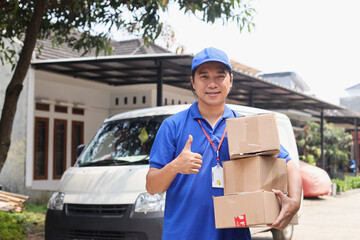 Young Delivery Man in Blue Uniform Carrying Pile of Cardboard Box and Showing Thumb-up In Front Of...