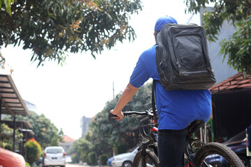 Rear view and close-up of deliveryman wearing blue uniform bring packages on bag riding bicycle to...