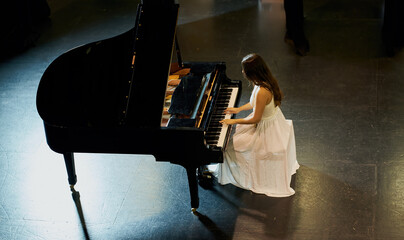 A woman with brown hair dressed in a white dress seen playing a black grand piano with the lid raised. View from above of the pianist
