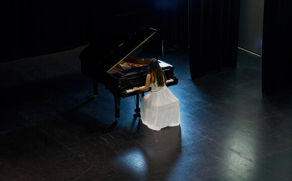 A woman with brown hair dressed in a white dress seen playing a black grand piano with the lid raised. View from above of the pianist