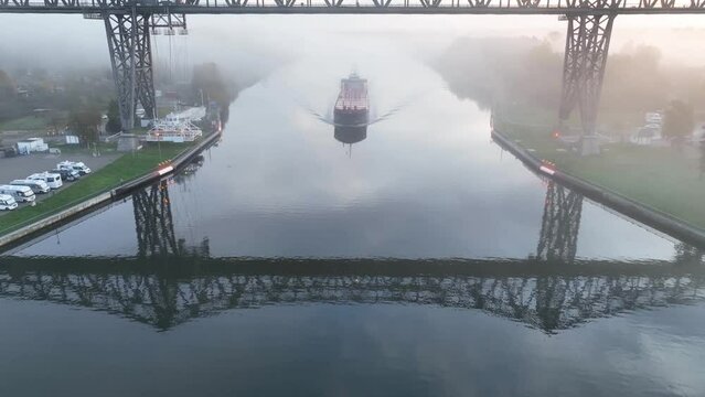 A cargo ship appearing from the morning mist and passing Rendsburg High Bridge railway viaduct with aerial transfer bridge over Kiel Canal. Drone footage of camping caravan area along Kiel Canal.