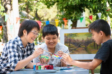 Asian middle-aged female art teacher is encouraging and explaining the process of watercolor painting to young asian boys during their art lesson in the school park, raising teens concept.