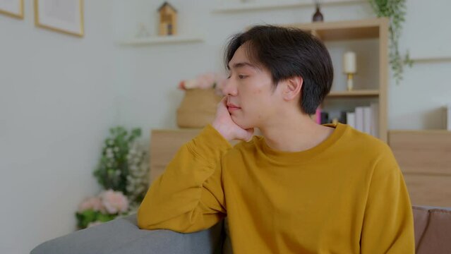 Thinking asian man sitting on sofa in house. stressed out about personal life, finances, Family, work and girlfriend. Stress that causes migraines or depression on couch.