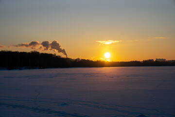 Sunset over the frozen surface of the river bay. The icy surface of the river covered with snow against the backdrop of the forest and the setting sun