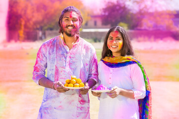 Portrait of happy young Indian couple wearing white kurta holding plate full of powder color celebrating holi festival at park outdoor, Face painted with colorful gulal