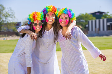 Group of young indian friends wearing white kurta and colorful hair wigs celebrating holi festival outdoor at park.