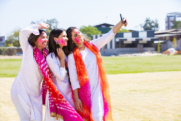 Naklejka premium Portrait of happy young indian female friends taking selfie picture with smart phone while celebrating holif festival together at park.