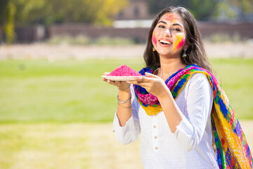 Portrait of smiling beautiful young Indian woman wearing white kurta dress holding pink powder color plate celebrating holi festival at park outdoor, Face painted with colorful gulal Copy space. - Powered by Adobe