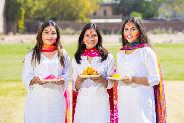 Group of happy young indian woman wearing white cloths celebrating holi festival at park while holding plates full of gulal, Girls having fun with colorful powder.