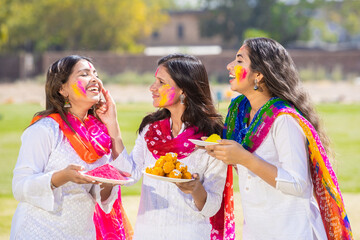 Obraz premium Group of happy young indian woman wearing white kurta dupatta outfit celebrating holi festival at park while holding plates full of gulal and laddoo sweet , woman having fun with colorful powder.