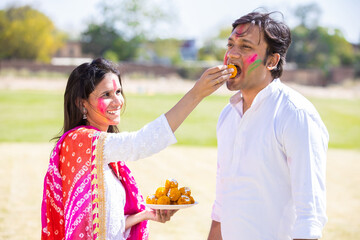 Obraz premium Happy young indian couple celebrating holi festival together while enjoying delicious laddu sweet.