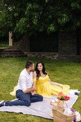 Beautiful couple sitting on a plaid on the grass. Elegant pregnant woman with husband waiting for baby. Future parents. Yeallow dress