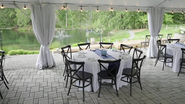 Wooden table decorated for a wedding banquet in a beautiful estate. There is a big tent and a lake with a fountain.
