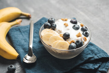 Granola cereal oatmeal with white yogurt, blueberries and banana fruits in a bowl on a blue napkin, grey background