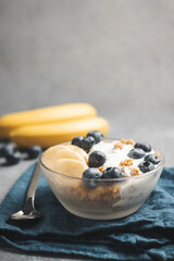 Granola cereal oatmeal with white yogurt, blueberries and banana fruits in a bowl on a blue napkin, grey background