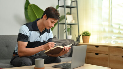 Millennial man using laptop nd making important notes on notepad, working online from home