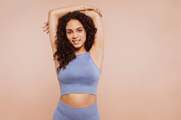 Portrait of a young beautiful multiracial curly-haired brunette woman wears sportswear, stretching arms at studio isolated over beige background. Fitness, healthy lifestyle concept.