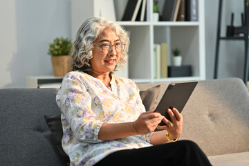 Smiling senior woman relaxing on comfortable sofa and browsing wireless Internet on tablet. Elderly technology concept