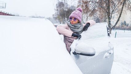 young woman with a hat, gloves and a scarf looking at a windshield covered with snow. High quality...
