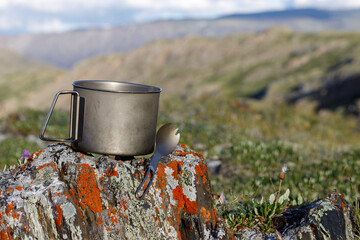Camping titanium mug with a spoon on the background of the Alpine valley. Objects in focus, background blurred.