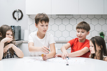 Happy little school friends play board game on paper map with dice and chips at kitchen table....