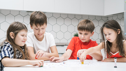 Funny little school friends play board game on paper map with dice and chips at kitchen table. Children throw dice and move figure. Home leisure, playtime. Entertaining interesting table game.