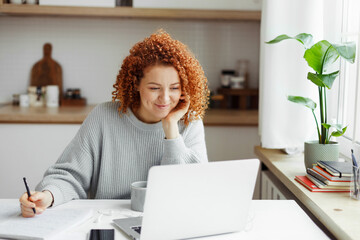 Cute girl with red curls watching online tutorial or lesson on laptop, smiling listening and looking attentively, making notes in copybook, sitting at kitchen table with smartphone and cup of coffee - obrazy, fototapety, plakaty
