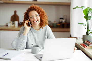 Young redhead accountant manager working from home at kitchen table, talking on phone in front of laptop, smiling, having nice conversation with ceo or bank support service about corporate account