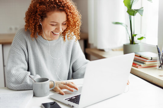 Student with red curls studying at home, having rest video-chatting with her mates on laptop, sitting at kitchen table with coffee cup and smartphone in wired earphones, laughing looking at screen