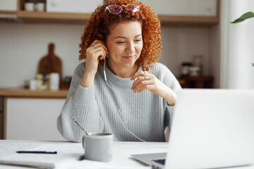 Portrait of redhead female with curly hair in sweater sitting in front of laptop at kitchen table with copybook, pen and cup of coffee, in wired headphones, watching webinar or online lesson - Powered by Adobe