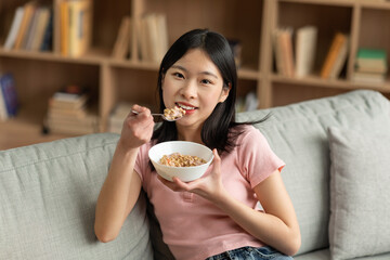 Balanced nutrition concept. Japanese lady eating granola or cereal with milk, sitting on comfy...