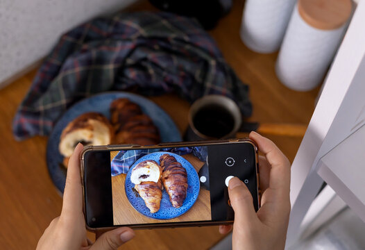 Woman taking picture of breakfast in cafe. Croissant, coffee with cream and jam on wooden table. Toned image, lifestyle and social media concept