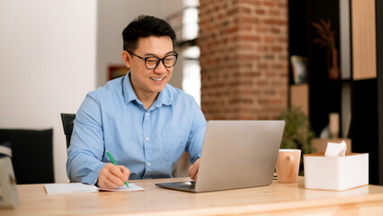 Positive middle aged asian man in glasses working on laptop and making notes, sitting at table in...