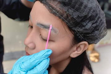 An esthetician applies numbing cream or topical anesthesia to a clients eyebrows prior to a...