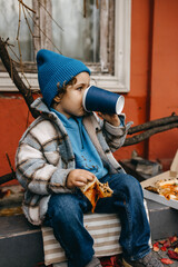 Little boy eating pizza and drinking soda, outdoors, sitting on city street.