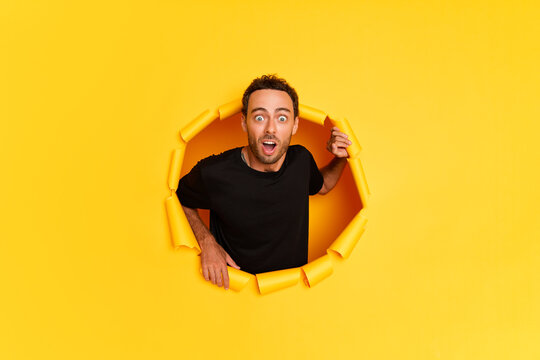 Astonished Caucasian Young Man In Black T-shirt With Wide Opened Mouth Gasps From Amazement Shocked From Something Poses In Torn Paper Wall. Advertisement Concept 